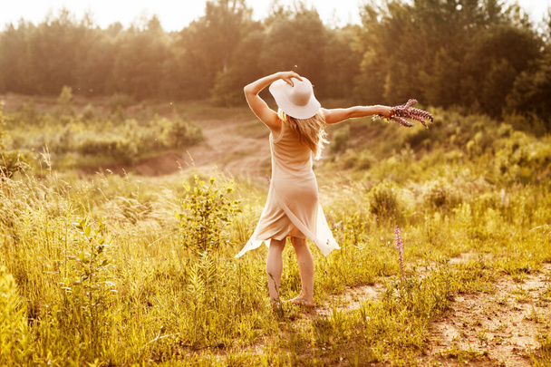 Молода дівчина в легкій сукні і капелюх з букетом в руках танцює на полі, дивлячись на захід сонця. Вид ззаду, простір для тексту
 - Фото, зображення