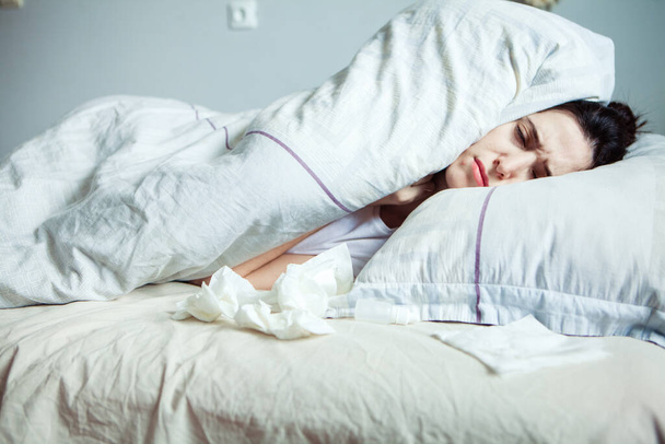 A young woman feels cold in a bed. She is trying to warm up under the blanket. Sick girl buries a drop of cold in the nose. Cold weather concept.Woman lying in bed and having fever  - Photo, image