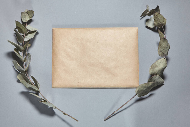 A brown craft envelope on gray background. Closed craft envelope, branch of eucalyptus on gray table. Invitation card mockup. Flat lay, top view, copy space, mocku - Photo, Image