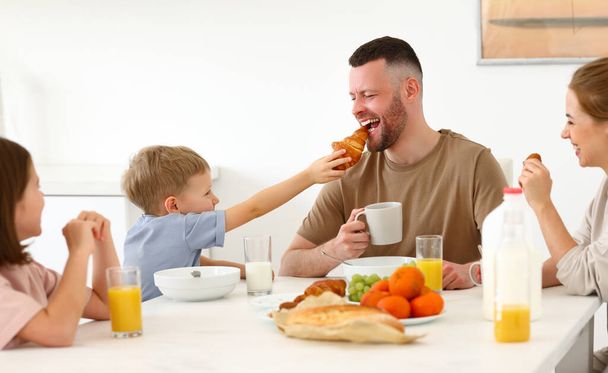 若い幸せな美しい家族は家で一緒に朝食を持っています。お父さん、お母さんと2人のかわいい子供たちは、午前中に健康的な食べ物を食べて、現代のキッチンに座って話して笑顔 - 写真・画像