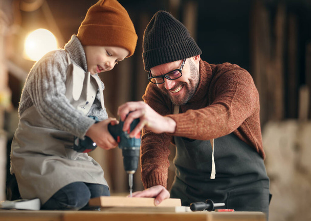 Ammattitaitoinen käsityöläinen opettaa pientä poikaa käyttämään poraa samalla kun tehdään yhdessä puinen lintutalo puusepänverstaassa - Valokuva, kuva