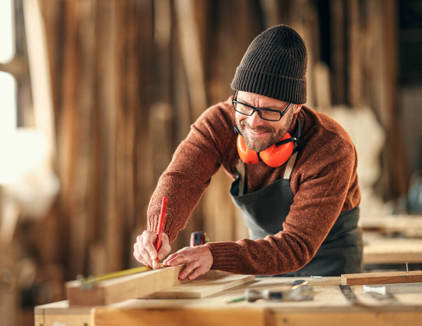 Skoncentrowany dorosły stolarz znakujący drewniany kawałek ołówkiem podczas pracy w pracowni stolarskiej - Zdjęcie, obraz