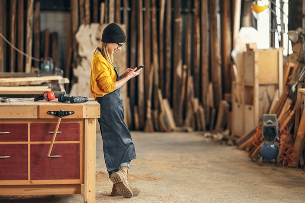 Full body side view of positive young female woodworker in the zástěra messaging on mobile phone while standing near workdesk in teinery workshop - Fotografie, Obrázek