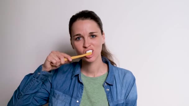 Fiatal aranyos fehér lány kék ingben mossa a fogát fehér háttér sárga fogkefével. A nő naponta gondoskodik a szájhigiéniáról. 4K felvétel kiváló minőségű. - Felvétel, videó