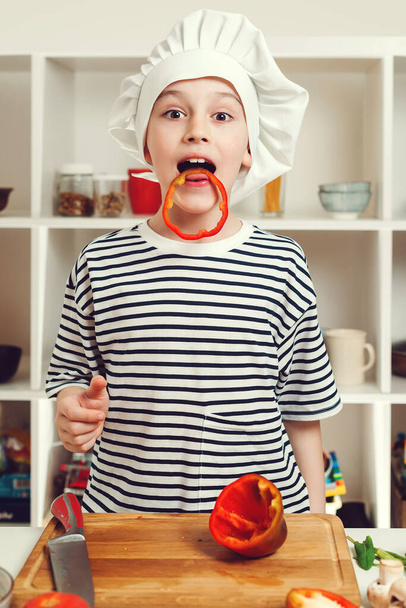 キッチンで楽しんでいるかわいい小さなシェフの肖像画。シェフの帽子をかぶった少年。子供は将来の職業を夢見ている。サラダ用のホームカットコショウでキッチンのシェフ。料理教室の少年. - 写真・画像