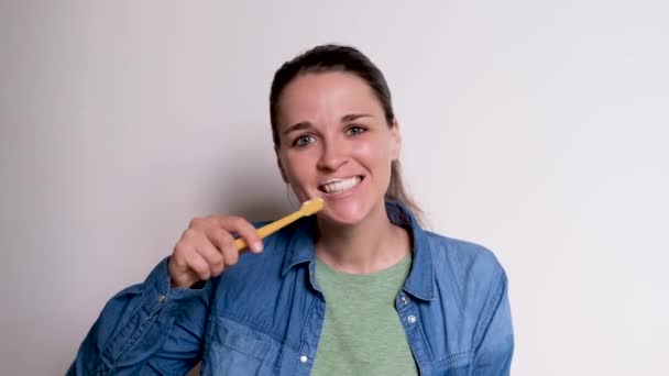 Mavi gömlekli genç beyaz kız dişlerini sarı diş fırçasıyla beyaz arka planda fırçalıyor. Kadınlar günlük olarak ağız hijyeniyle ilgilenirler. Yüksek kalitede 4K görüntü. - Video, Çekim
