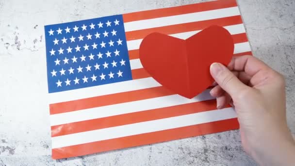 Λατρεύω τις ΗΠΑ. Πλάνα χεριών που κρατούν ένα κόκκινο σύμβολο της καρδιάς στο φόντο της αμερικανικής σημαίας.   - Πλάνα, βίντεο