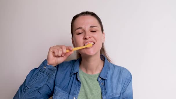 Jeune fille blanche mignonne en chemise bleue se brosse les dents sur fond blanc avec une brosse à dents jaune. Femme prend soin de l'hygiène buccodentaire sur une base quotidienne. Images 4K de haute qualité. - Séquence, vidéo
