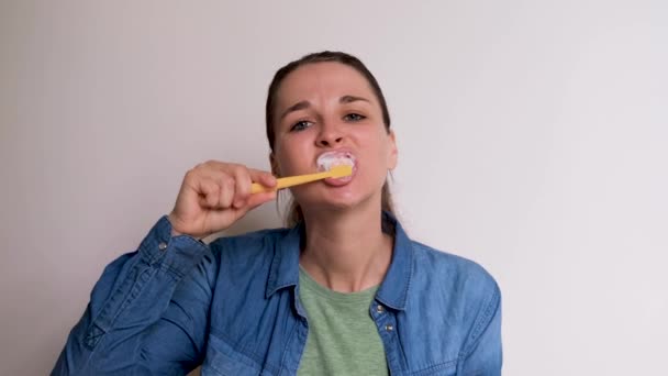 Menina branca bonito nova na camisa azul está escovando os dentes no fundo branco com escova de dentes amarela. A mulher cuida da higiene bucal diariamente. Imagens 4K de alta qualidade. - Filmagem, Vídeo