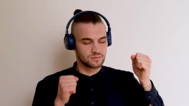 Young handsome Caucasian European man with beard and ponytail in shirt is dancing on white background while listening to music through large headphones. 4K footage of dancing man. - Footage, Video