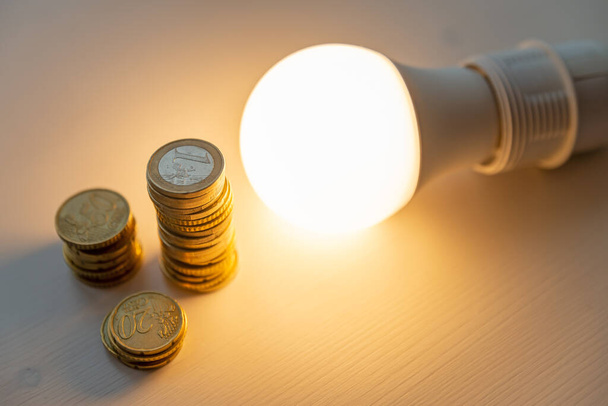 Λαμπτήρας LED αναμμένος, με νομίσματα δίπλα του, σε λευκή επιφάνεια. Κόστος της ενέργειας, αύξηση των τιμολογίων ενέργειας  - Φωτογραφία, εικόνα