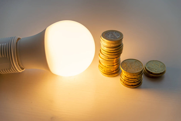 Λαμπτήρας LED αναμμένος, με νομίσματα δίπλα του, σε λευκή επιφάνεια. Κόστος της ενέργειας, αύξηση των τιμολογίων ενέργειας  - Φωτογραφία, εικόνα