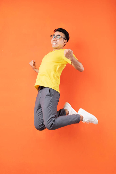 黄色のTシャツのアジア人男性がオレンジの背景にジャンプ - 写真・画像