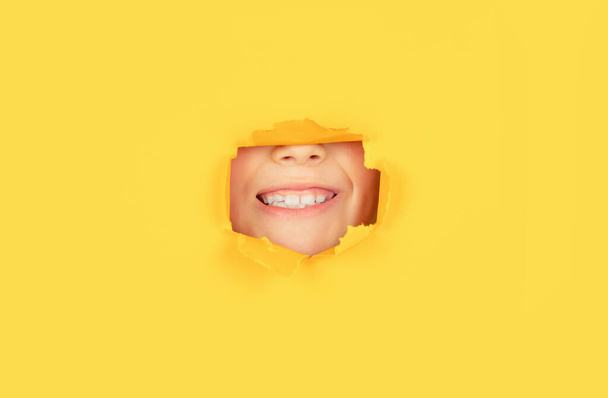 Positive child with toothy pleasant smile on face, keeps through torn hole in yellow paper. Breaking paper background. Emotions concept. Kid with toothy smile shows face in paper hole - Foto, Imagem