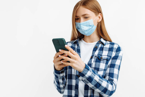 Young woman in a medical mask, uses a mobile phone, on an isolated white background, Coronavirus pandemic - 写真・画像