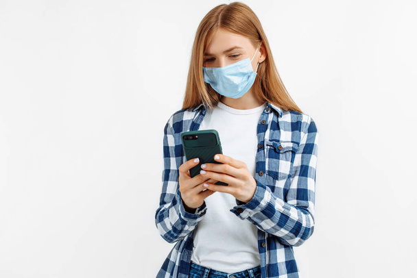 Portrait of a smiling young woman, wearing a medical mask for protection from covid 19, holding a smartphone on an isolated white background - Foto, Bild