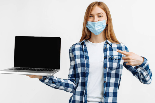 Mujer joven feliz, en máscara médica, mostrando la pantalla del ordenador portátil en blanco sobre fondo blanco - Foto, Imagen