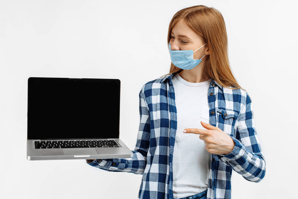 Happy young woman, in medical mask, showing blank laptop screen on white background - Foto, afbeelding