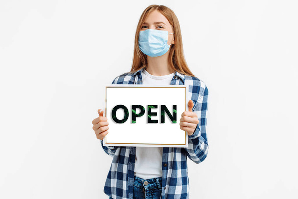 Young woman in protective mask holding a sign with a closed inscription, on an isolated white background, closing due to a pandemic - Foto, afbeelding