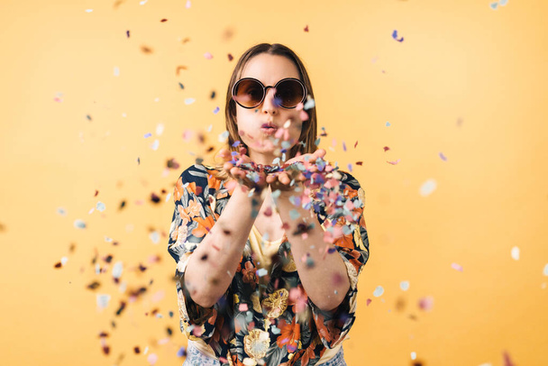 young woman in a flowered shirt and big round sunglasses blowing confetti on a yellow background - Photo, image