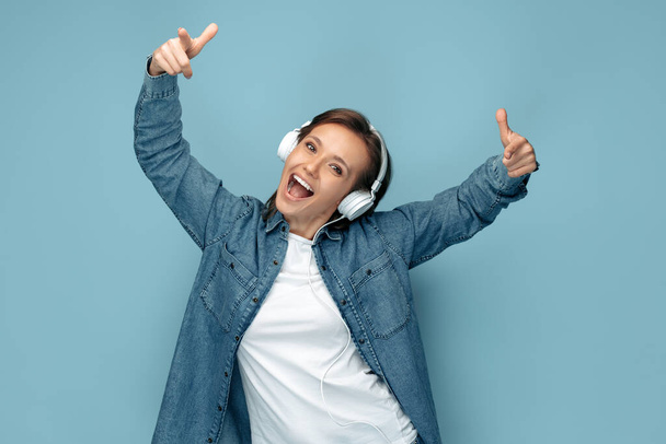 Happy young woman in jeans shirt wearing headphones dancing over blue background. - Fotoğraf, Görsel