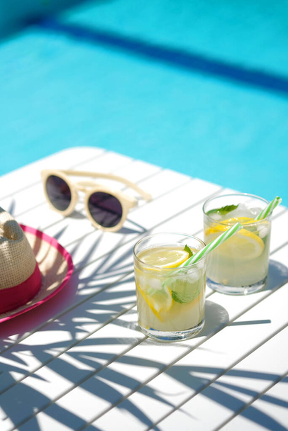 λεμονάδα σε ποτήρια με φέτα λεμονιού, ψάθινο καπέλο και γυαλιά ηλίου σε λευκό τραπέζι, άκρη της πισίνας με τυρκουάζ φόντο μπλε νερό, φως και σκιές, κλίση και το καλοκαίρι έννοια - Φωτογραφία, εικόνα