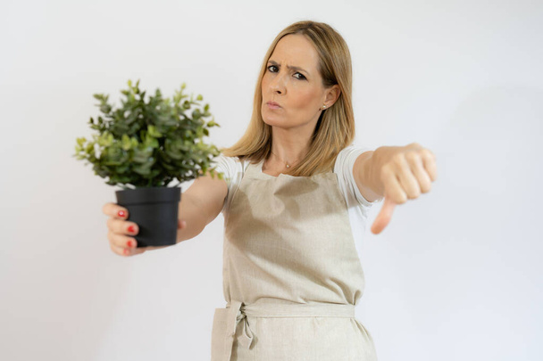 Mujer joven sosteniendo una planta con expresión triste con el pulgar hacia abajo aislado sobre fondo blanco. - Foto, Imagen