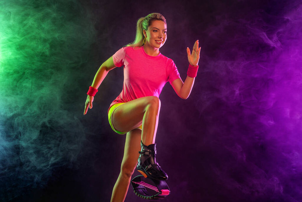 A Strong athletic, female runner on the black bacground wearing a tight, fitness outfit. Sports girl in shoes for kangoo jumping. Blonde woman and fitness athlete isolated on color background. - Foto, immagini
