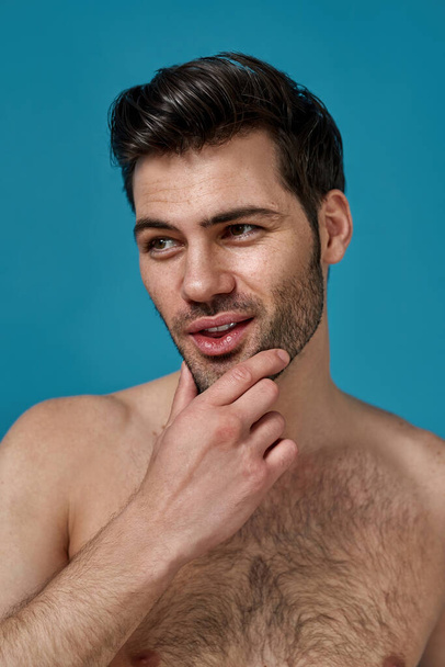Portrait of handsome naked brunette guy with fresh skin looking aside, touching his chin while posing isolated over blue background - Valokuva, kuva