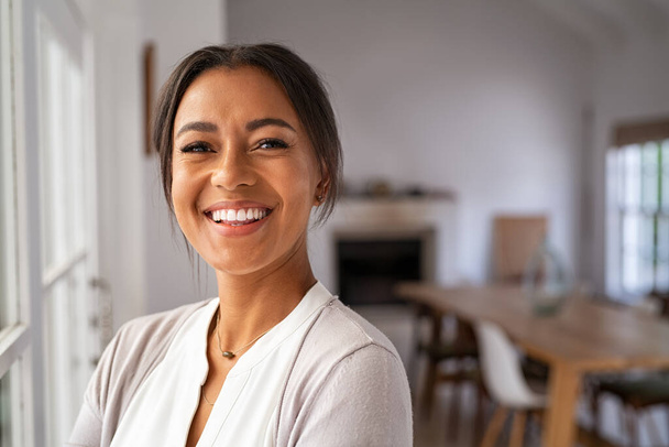 Portrait of a beautiful african woman smiling while looking at camera. Mature black woman in casual standing near window at home. Close up face of laughing cheerful lady near window with copy space. - Foto, Bild