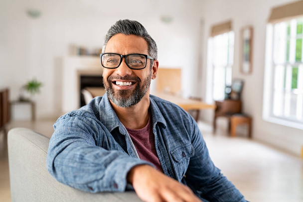 Happy mature middle eastern man wearing eyeglasses with beard sitting on couch at home. Portrait of indian man relaxing at home and looking away with big smile. Handsome mid adult guy with specs thinking about his future. - Zdjęcie, obraz