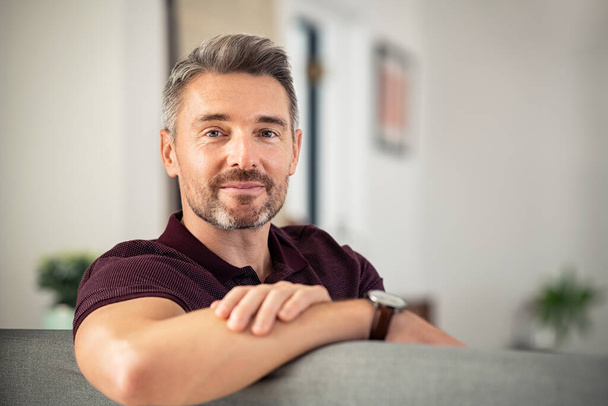Portrait of handsome mature man relaxing on couch at home while looking at camera. Confident mid adult man resting while sitting on sofa in living room with copy space. Reliable businessman looking at camera with satisfaction. - Фото, изображение