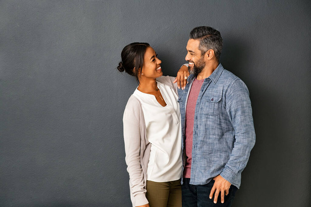 Happy mature indian couple embracing and looking at each other against gray background. Mid adult multiethnic couple in love standing against grey wall. Middle eastern man fall in love of his beautiful hispanic girlfriend. - Foto, imagen