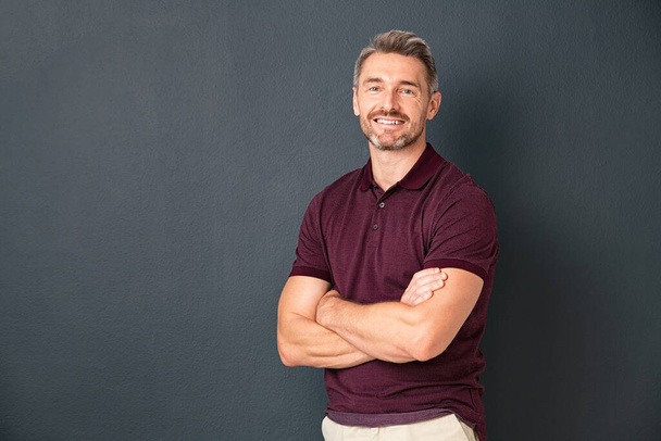 Portrait of smiling mature man standing on gray wall with folded arms. Successful mid adult man with folded arms standing over grey background while looking at camera. Happy healthy guy on grey wall with copy space. - Fotografie, Obrázek