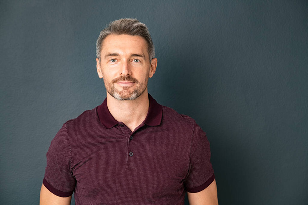 Portrait of middle aged man in casual clothing standing isolated on grey background. Successful mature man standing against gray wall and looking at camera with smile. Confident reliable guy standing with copy space against wall. - Фото, зображення