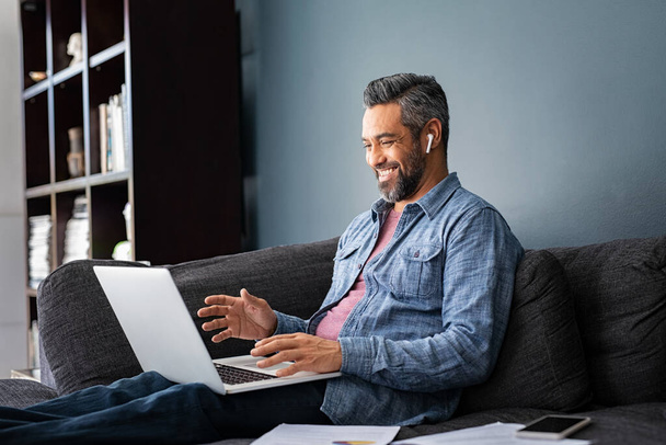 Happy mature indian man sitting on couch while doing a video call with laptop. Mixed race businessman sitting on sofa and making a video conference with wireless earphones while working from home, new normal concept. Smiling mid adult ethnic business - Photo, image