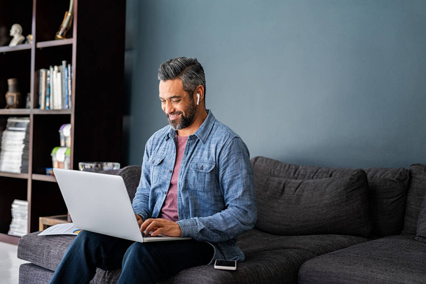 Successful mature indian businessman sitting on couch typing on laptop with wireless earphones. Mixed race businessman sitting on couch while working from home during video call. Happy multiethnic business man in casual clothing working on computer.  - Fotografie, Obrázek