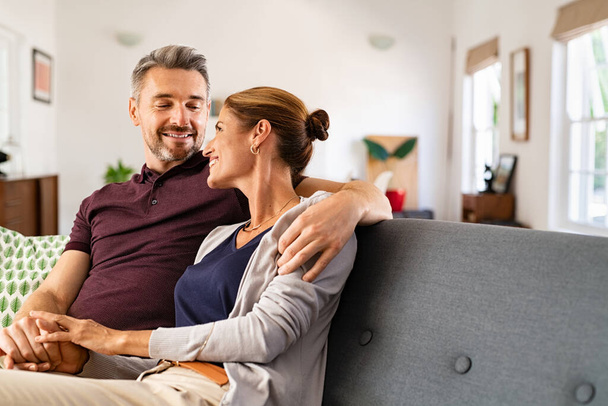 Mid adult couple embracing and relaxing on couch at home while holding hands. Romantic husband and beautiful wife hugging and looking at each other on sofa. Middle aged man relaxing at home with smiling woman. - Fotoğraf, Görsel