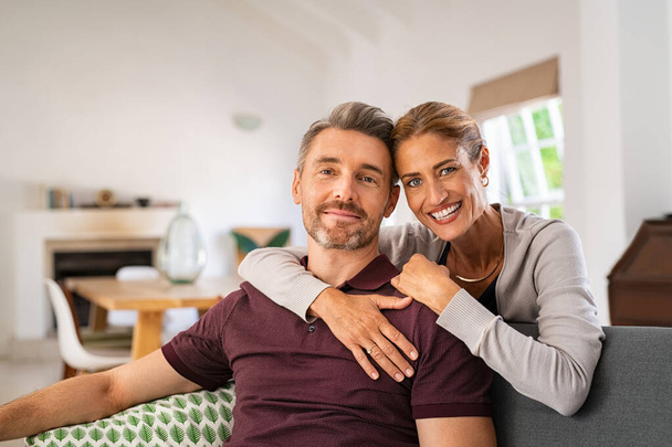 Middle aged couple embracing on sofa while looking at camera. Mature happy woman embracing her husband from behind while relaxing at home. Portrait of smiling wife and man loving in perfect harmony. - Photo, image