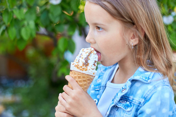 Słodka dziewczyna z włoskim rożkiem lodowym uśmiechnięta i patrząca w kamerę podczas odpoczynku w parku w letni dzień, dziecko cieszące się lodami na świeżym powietrzu, szczęśliwe wakacje, lato - Zdjęcie, obraz