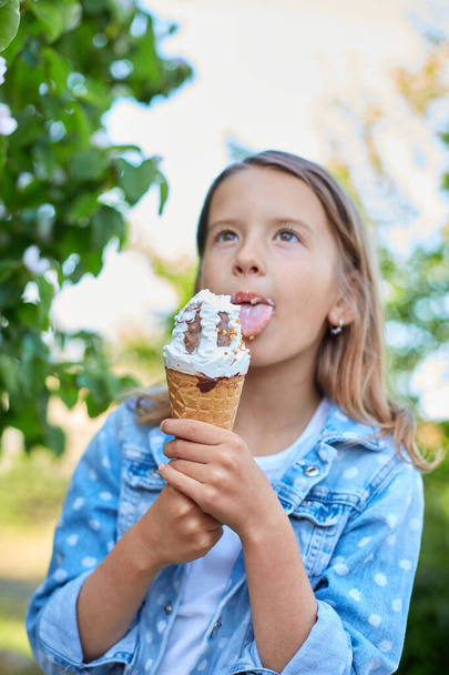 Χαριτωμένο κορίτσι με ιταλικό παγωτό χωνάκι χαμογελώντας και κοιτάζοντας την κάμερα ενώ ξεκουράζεται στο πάρκο την καλοκαιρινή μέρα, το παιδί απολαμβάνει το παγωτό εξωτερική, χαρούμενες διακοπές, καλοκαίρι - Φωτογραφία, εικόνα
