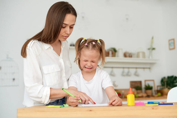 Kind mother embracing and comforting small crying daughter having difficulties with homework while sitting at table and doing exercise in copybook in kitchen. Help with homework - Photo, Image