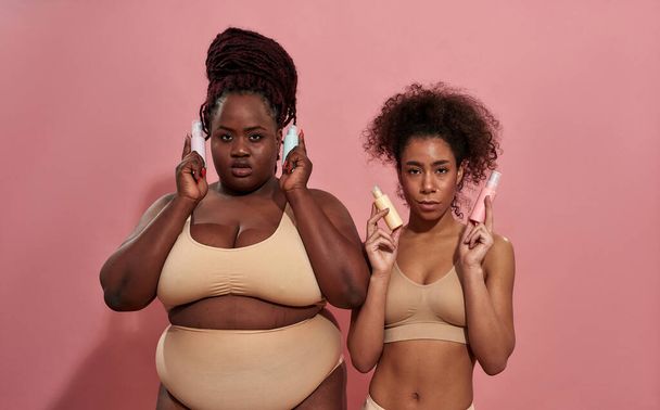 Couple of young afro girlfriends seductively watching in camera and holding cosmetics - Fotoğraf, Görsel