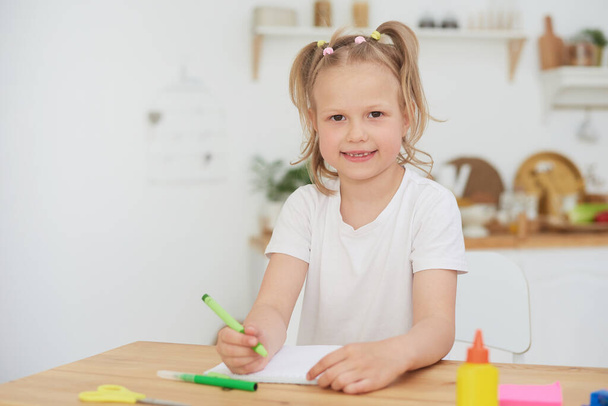 Portrait of cute girl smiling at camera while doing homework or tests in cozy home interior. Homeschooled child during coronavirus - Photo, Image
