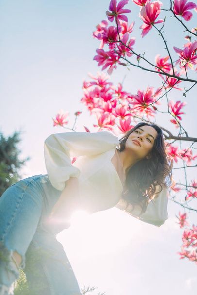 Young woman poses near blooming magnolia tree in spring garden against sky background. Backlight. - Photo, Image