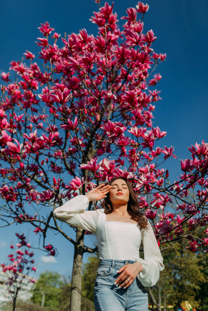 若い女性が空を背景に春の庭でマグノリアの木を咲かせる近くに立つ. - 写真・画像
