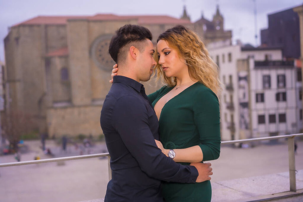Fashionable pose of a Caucasian loving couple in a blue suit and green dress, caressing each other in the city and looking at each other about to kiss - Fotó, kép