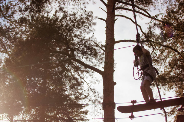 Ein Teenager fliegt mit einem Skateboard in einem Seilpark mit Extremsportgeräten. Ein Hochseilgarten hoch oben zwischen den Bäumen. Abenteuer-Konzept. Schulkinder im Urlaub. Eine Jungensilhouette im Kontrast. - Foto, Bild