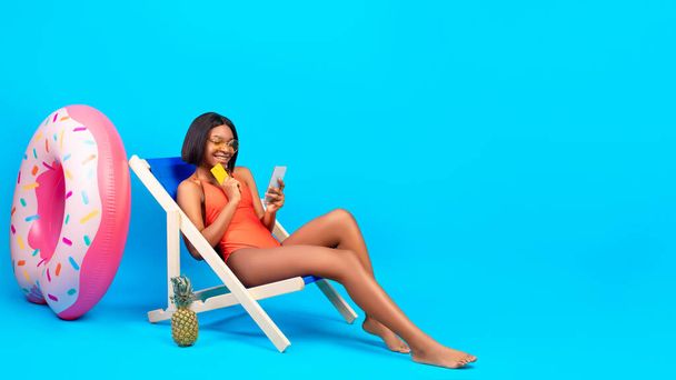 Donna nera spensierata in possesso di carta di credito e smartphone, shopping online sulla sedia a sdraio su sfondo blu - Foto, immagini