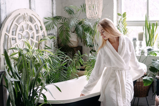 Beauty concept. Charming young woman in white bathrobe draws water into the bath. Female standing near freestanding tub, spending morning in bathroom with houseplants and soft daylight - 写真・画像